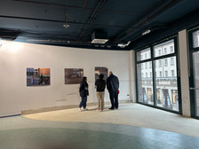 Drei Personen stehen vor drei Kunstwerken, die an einer weißen Wand in einem hell erleuchteten Galerieraum mit großen Fenstern mit Blick auf eine Stadtstraße ausgestellt sind.