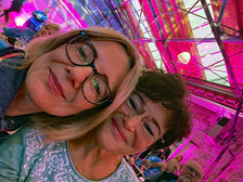Zwei Frauen lächeln eng beieinander in einem lebhaften, rosa beleuchteten Innenraum mit einer Glasdecke. Im Hintergrund sitzen weitere Personen, was auf ein gesellschaftliches Treffen oder eine Veranstaltung hindeutet.