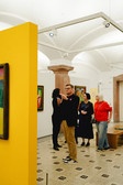 Eine Gruppe von Menschen betrachtet und diskutiert Kunstwerke in einer hell erleuchteten Kunstgalerie mit weißen Wänden und gewölbten Decken. Ein Mann im Vordergrund gestikuliert, während er ein Getränk in der Hand hält. An den Wänden hängen Gemälde und in der Nähe steht ein auffälliger gelber Raumteiler.