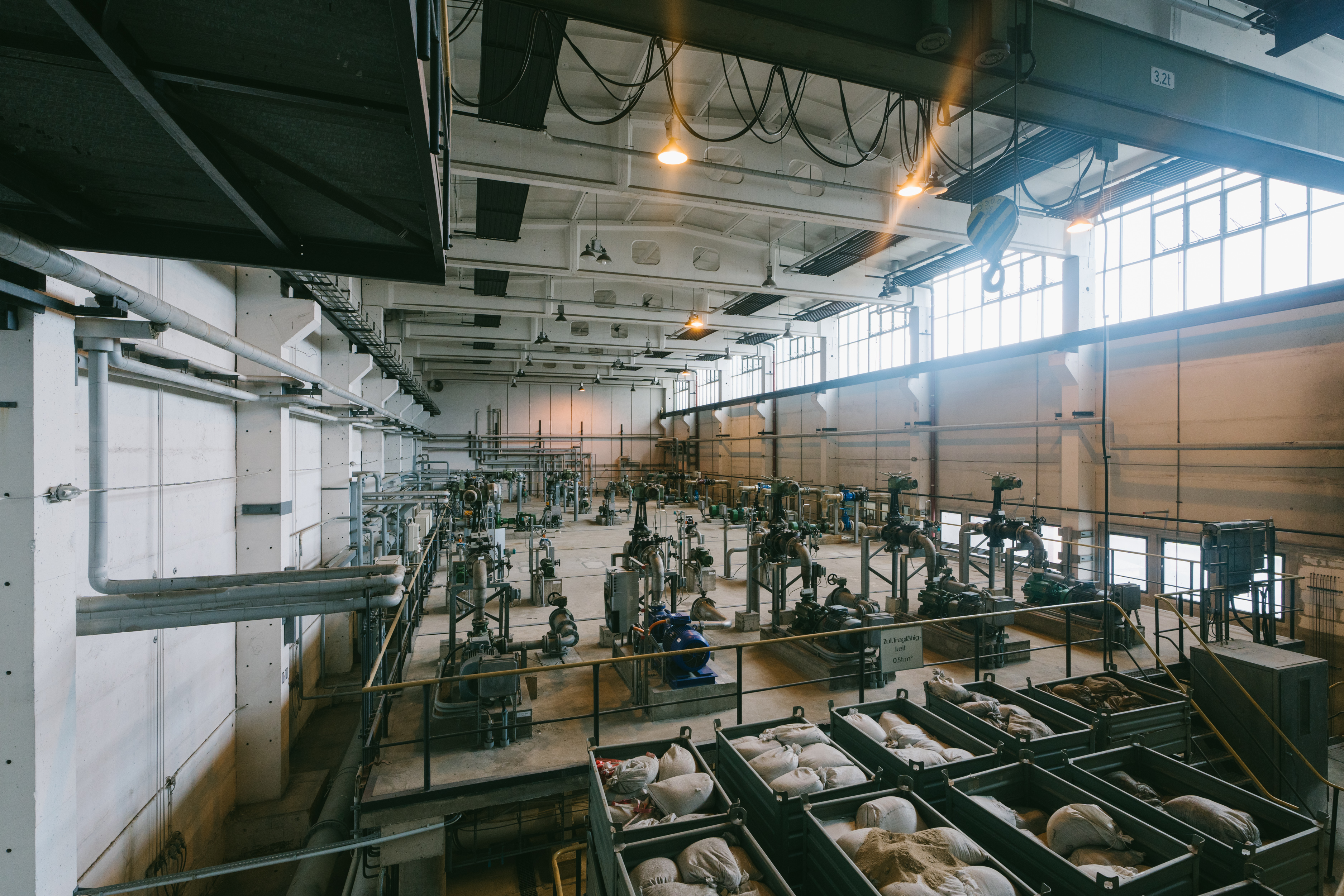 Eine weitläufige Industrieanlage mit hohen Decken, großen Deckenlampen und zahlreichen großen Rohren und Maschinen. Im Vordergrund sind mehrere große Behälter mit weißen Säcken zu sehen. Sonnenlicht fällt durch hohe Fenster auf der rechten Seite ein.