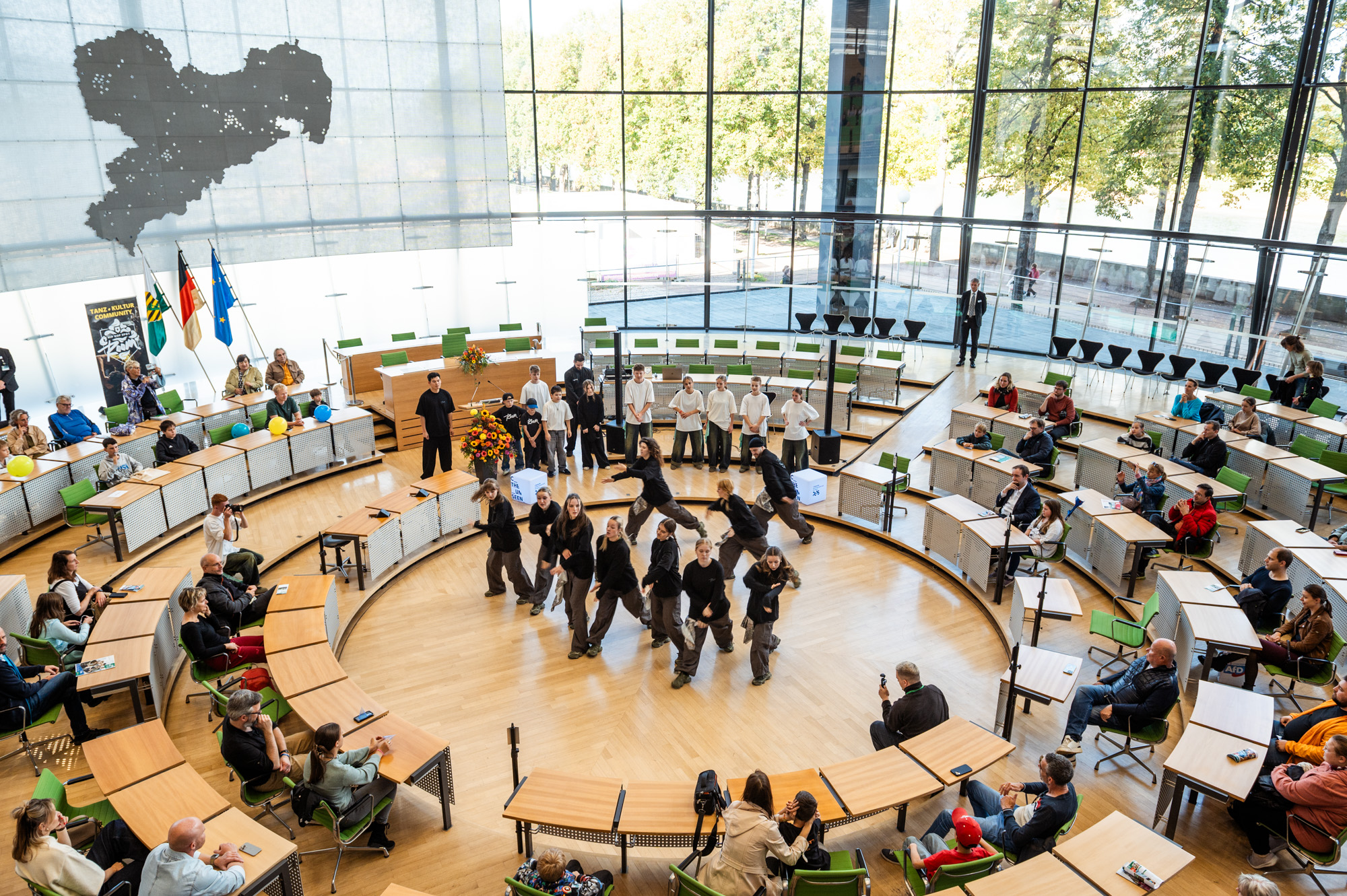Eine Gruppe führt in der Mitte eines runden, modernen Auditoriums mit großen Fenstern einen Tanz auf. Die Leute sitzen herum und schauen zu, und auf der linken Seite sind Fahnen aufgestellt. Draußen sind Bäume und Sonnenlicht zu sehen.