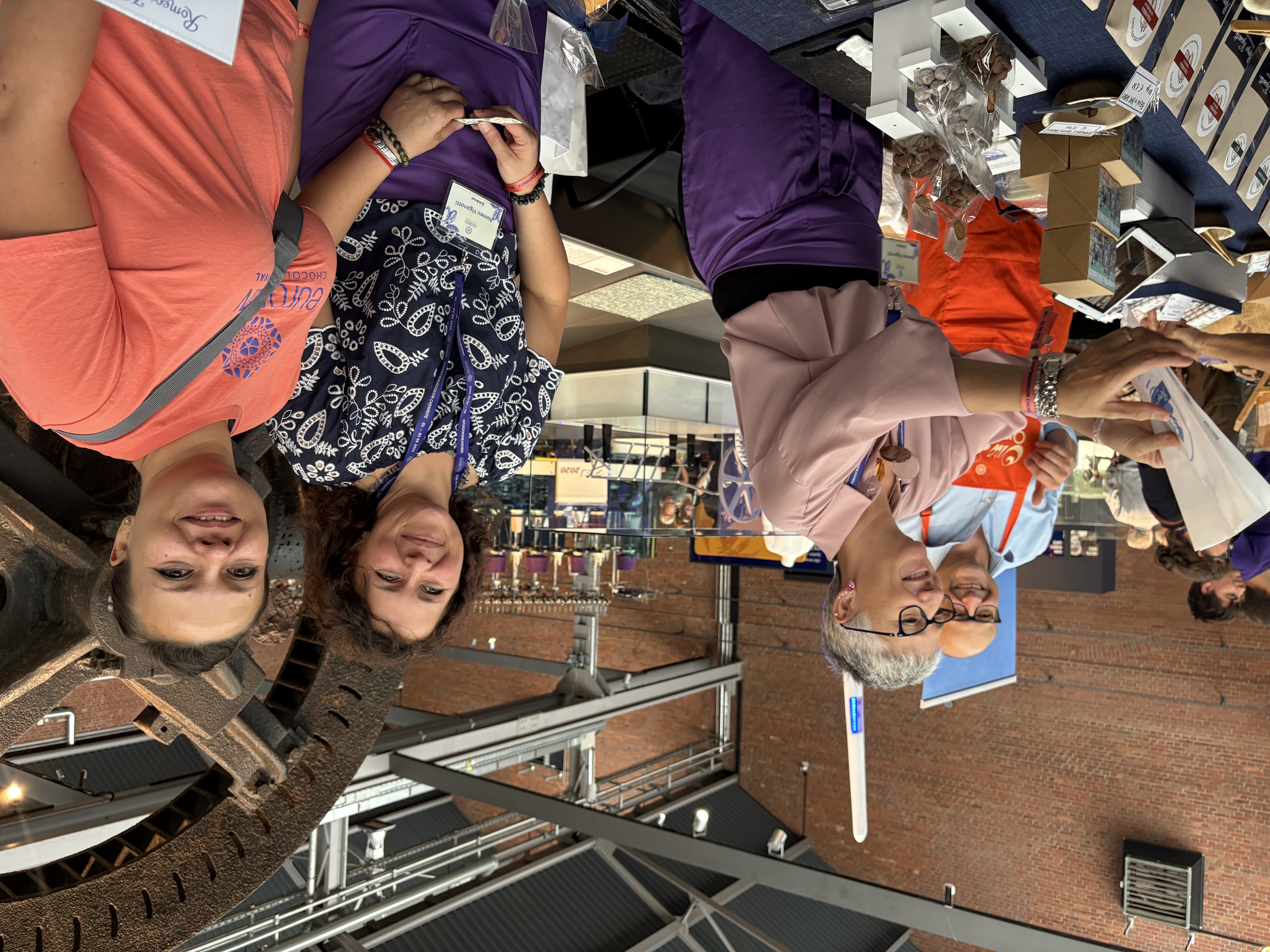 Vier lächelnde Menschen stehen hinter einem Tisch mit verschiedenen Pralinen und Leckereien auf einem Markt oder einer Veranstaltung in einer Halle, mit Industriemaschinen und Backsteinmauern im Hintergrund. Eine Frau reicht einem Kunden einen Artikel.
