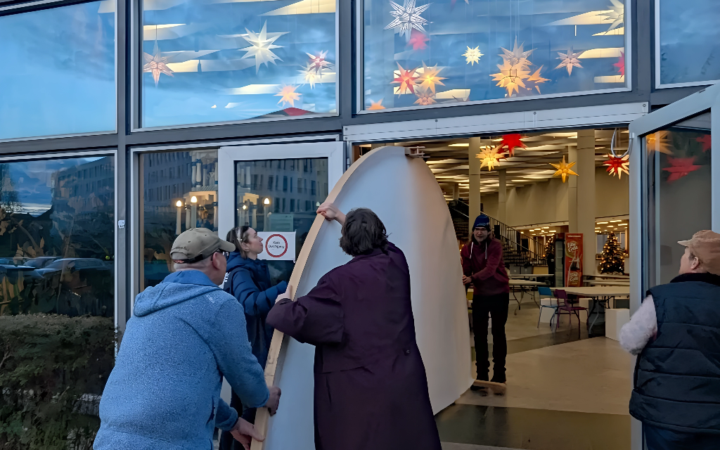 Vier Personen arbeiten zusammen, um ein großes, gekrümmtes Objekt durch doppelte Glastüren in ein hell erleuchtetes Gebäude zu bringen, das mit bunten sternförmigen Lichtern und einem Weihnachtsbaum im Inneren geschmückt ist.
