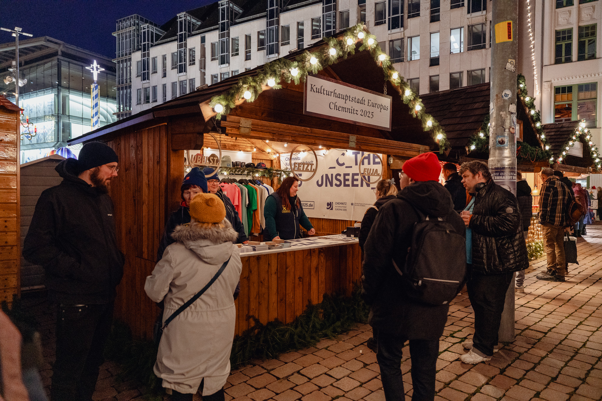 Menschen versammeln sich um einen festlichen Holzstand auf einem Nachtmarkt in Chemnitz, der mit Lichterketten geschmückt ist. Auf dem Schild oben steht "Kulturhauptstadt Europas Chemnitz 2025". Einige stöbern in den Angeboten, während andere sich in der Nähe unterhalten. Im Hintergrund sind Gebäude zu sehen.